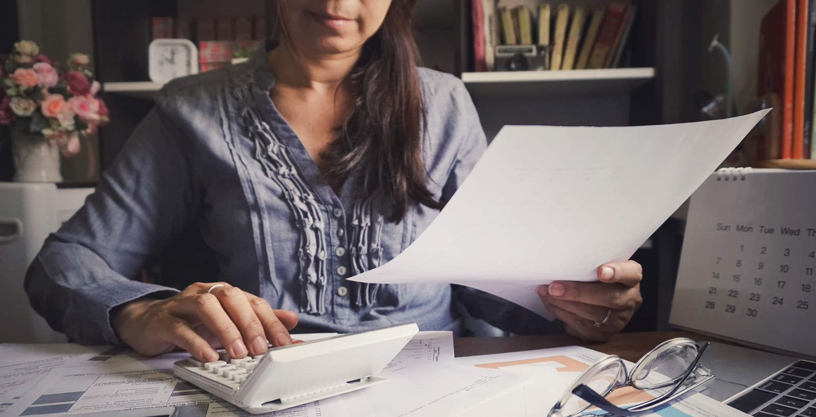 A woman holds a document while typing on a calculator