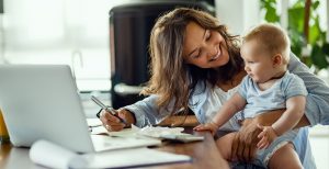 Mother smiles, holding her baby on her lap while doing paperwork in front of a laptop
