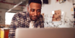 A man in a small workshop smiles while working at his laptop