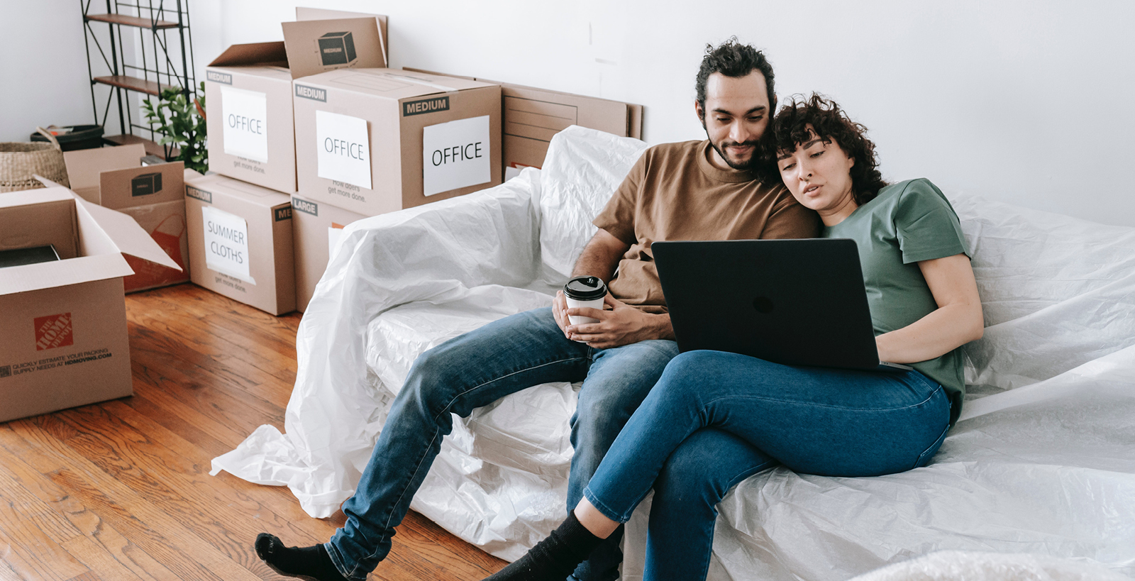 A young couple sits on a couch using a laptop, surrounded by unpacked moving boxes