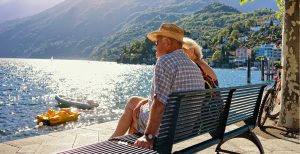 Elderly couple sits on a bench overlooking a bay with lush hills in the background.