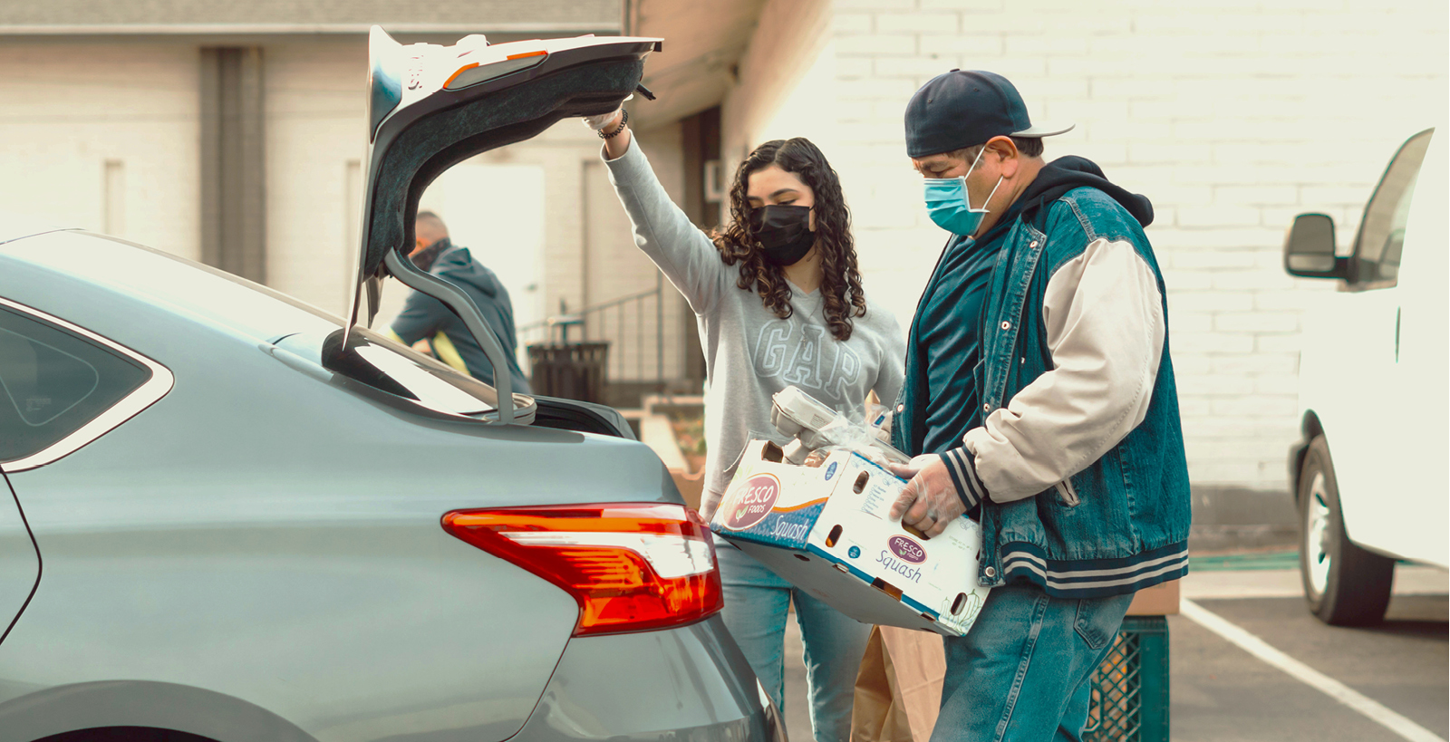 Masked man and woman load boxes into the trunk of a gray sedan