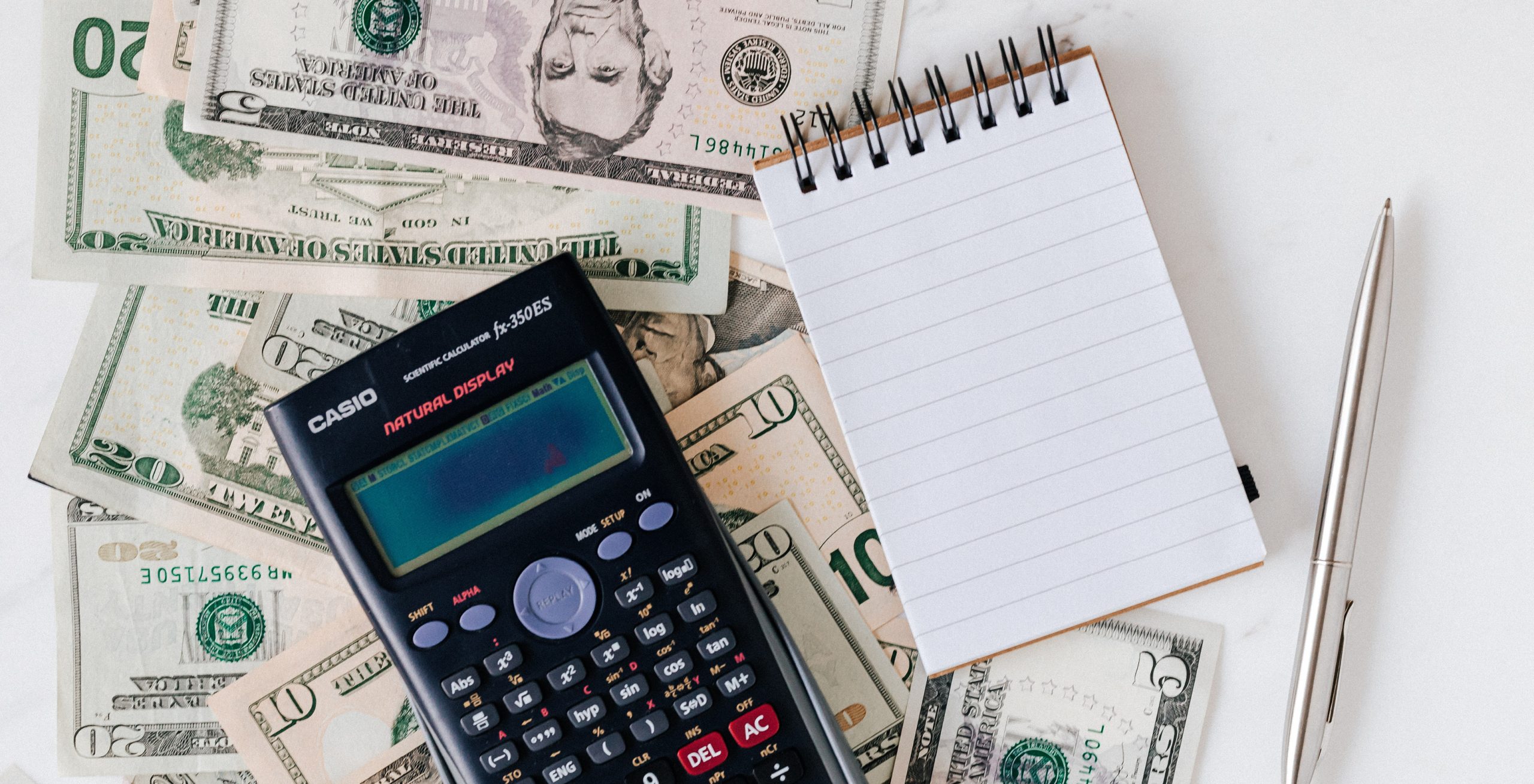 U.S. Dollars, a calculator, a small notebook, and a pen piled on a table.