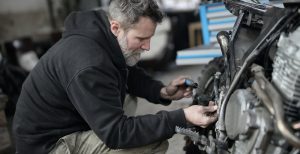 A man with a gray beard kneels to make repairs on a motorcycle