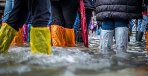A crowd of people, seen from the waist down, wearing plastic boot covers while standing in floor water up to their shins.