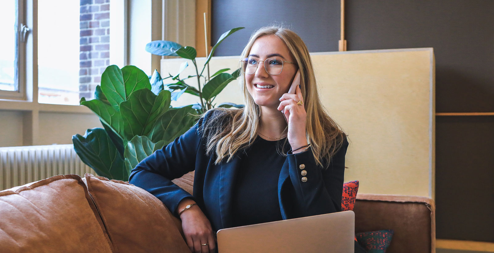 A blonde woman in a blazer smiles whiles on the phone. She's sitting on a brown suede couch with a silver laptop open on her lap.