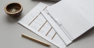 A budget sheet and financial tracking planner lay open on a gray desk beside a rose-gold pen and small wooden bowl of paperclips.