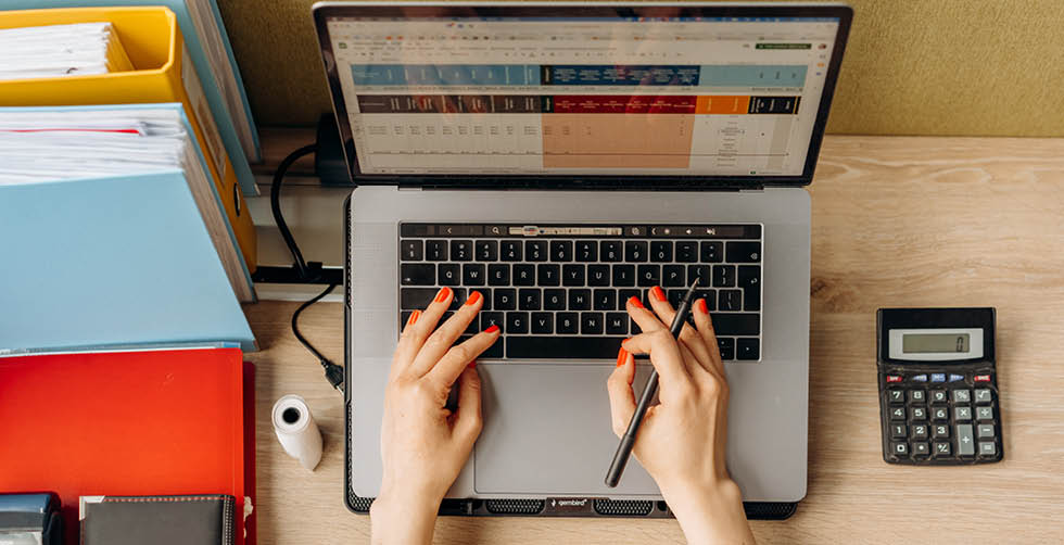 Hands with red nail polish type at a laptop featuring a spreadsheet. The surrounding desk has folders, binders, and a calculator.