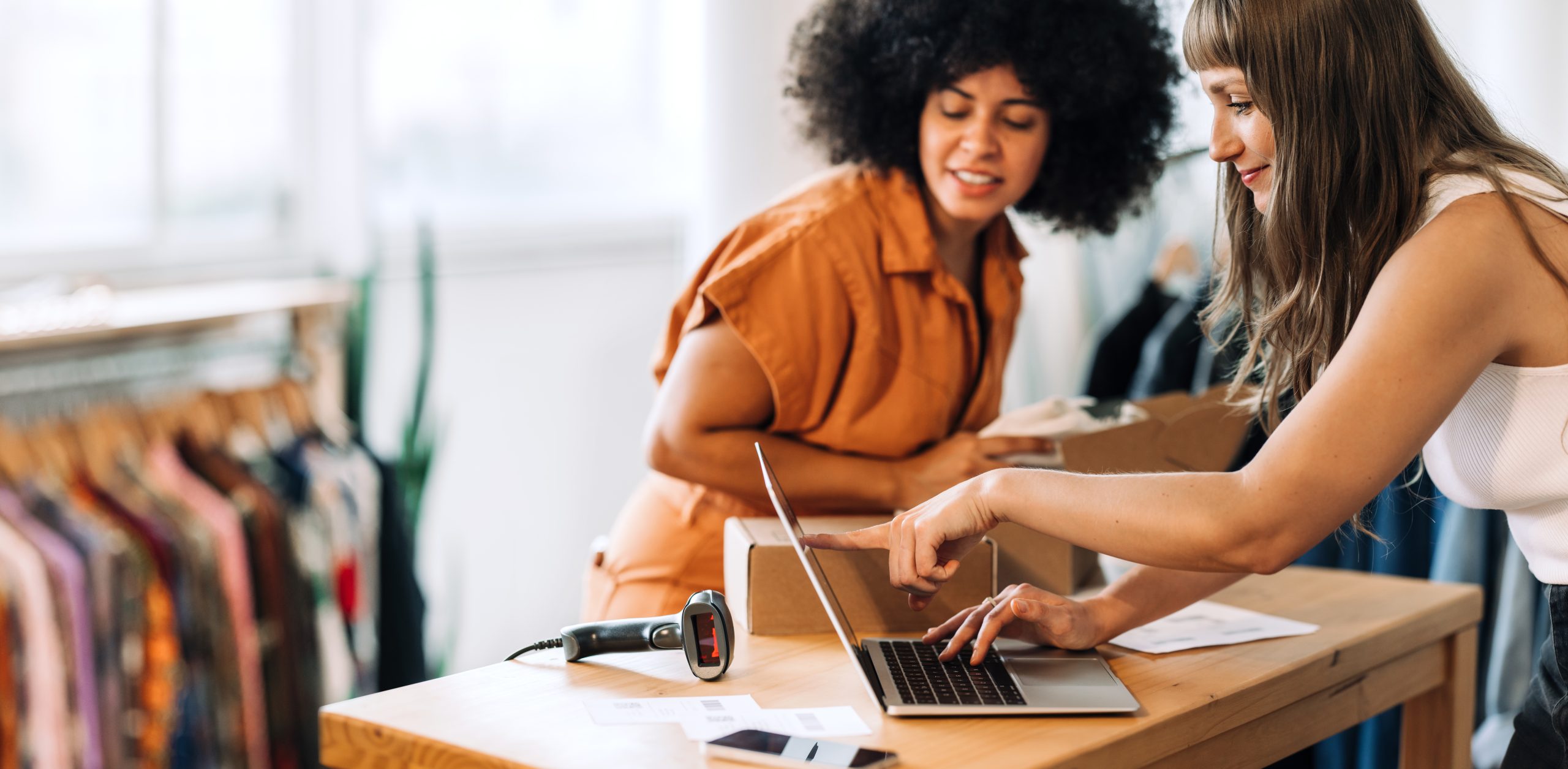 Clothing store owners having a discussion while preparing online orders for shipping. Two young women using a laptop together in a thrift store. Female entrepreneurs running an e-commerce small business.